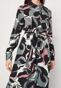 Šaty s dlouhým rukávem mají květinový vzor v černé, zelené a růžové barvě. Mají zapínání na knoflíky vpředu, límec a textilní pásek v pase.