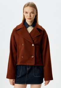 Giacca cropped doppio petto marrone in misto lana. Presenta ampi rever, due tasche anteriori e grandi bottoni marroni.