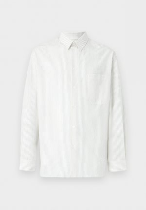 Young man with curly hair wearing a white shirt over a white tee, dark blue jeans, and brown sandals, standing against a plain light background.