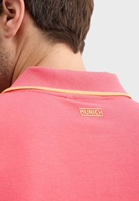 Ružový polo tričko s textúrovaným dizajnom, s oranžovým vyšitým logom "MUNICH" na golieri a oranžovými prešívacími detailmi.