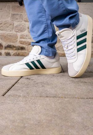 White sneakers with green three-stripe design, smooth leather upper, rounded toe, and contrasting grey suede toe cap. Rubber sole.