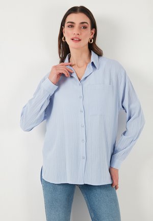 Light blue button-up shirt made of textured fabric, featuring a collar, long sleeves, and a chest pocket. Paired with denim bottoms.