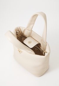 Sac à main en cuir beige doux avec des anses supérieures, rabat magnétique ouvert, plaque logo en ton or et doublure en tissu à l'intérieur.