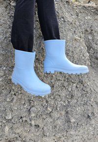 Lichtblauwe rubberlaarzen met een gladde textuur, ronde neus en robuuste zool; gedragen door een persoon die op ruw terrein staat.