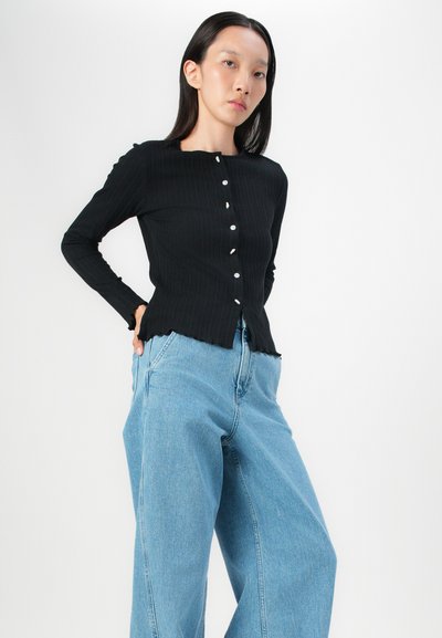 Zwarte geribbelde langwerpige top met ronde halslijn, knoopsluiting en gegolfde zoom, gecombineerd met lichtblauwe wijde jeans.