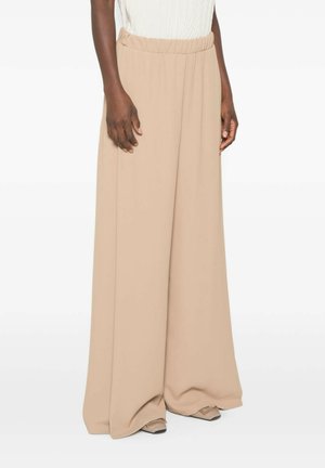 Persona che indossa pantaloni beige a vita alta e gamba larga con cintura elastica e scarpe beige appuntite su uno sfondo semplice.