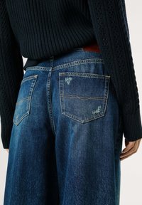 Persona que lleva un suéter de punto azul oscuro y unos pantalones vaqueros de pierna ancha de tiro alto, con bolsillos traseros y sutiles detalles desgastados.