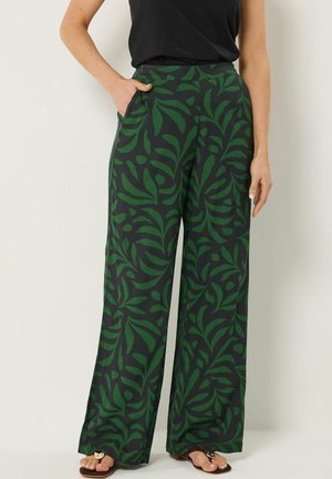 Femme portant un pantalon à jambes larges avec un motif feuille vert foncé, un haut noir et des sandales marron, debout avec une main dans la poche.