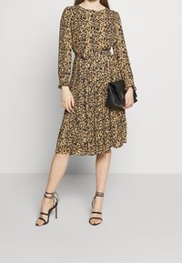 Vestido midi de estampado de leopardo con mangas largas, falda plisada y lazo en la cintura. Combinado con sandalias de tacón alto negras con tiras y un clutch negro.