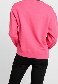 Różowa bluza z ribbowym dekoltem i mankietami. Charakteryzuje się luźnym krojem i gładką fakturą. Noszona z czarnymi dopasowanymi spodniami.