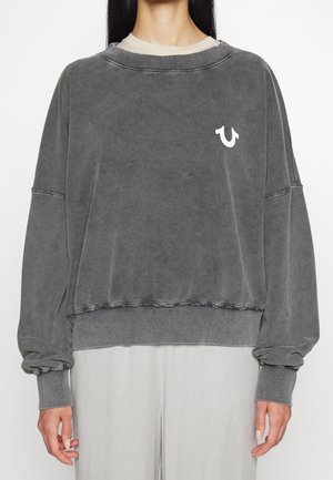 Kobieta w oversize wyblakłej ciemnoszarej bluzie z białym logo na lewej piersi i jasnoszarych spodniach, stojąca na białym tle.