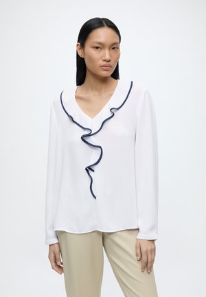 Femme portant un chemisier blanc à manches longues avec un volant bleu marine sur le devant et un pantalon beige, debout devant un fond uni.