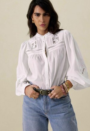 White button-up blouse with intricate lace detailing on the yoke and sleeves, paired with high-waisted blue denim jeans and a belt.