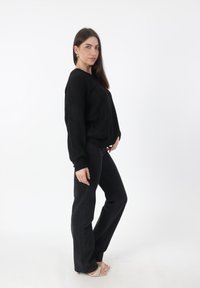 Zwarte gebreide trui met lange mouwen, gecombineerd met zwarte flared jeans. Model draagt beige strappy hakken. Effen witte achtergrond.
