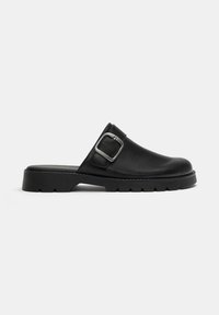 Chaussure noire en cuir à enfiler avec une semelle épaisse et une grande boucle argentée sur le côté, montrée en profil gauche sur fond blanc.