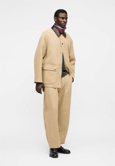 Άνδρας που φοράει μπεζ μπουφάν και φαρδιά παντελόνια, μαύρο πουκάμισο, πολύχρωμο φουλάρι στον λαιμό και μαύρα παπούτσια, στέκεται με το χέρι στην τσέπη.