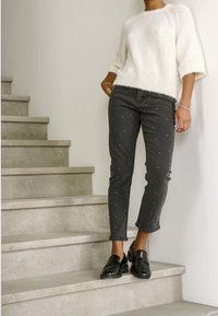 Personne portant un pull en laine blanc, un jean noir court avec de petits points blancs et des mocassins noirs, debout sur un escalier gris clair.