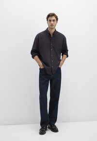 Dunkles Hemd mit aufgerollten Ärmeln, kombiniert mit locker sitzenden dunklen Jeans und schwarzen Schuhen, vor einem weißen Hintergrund.