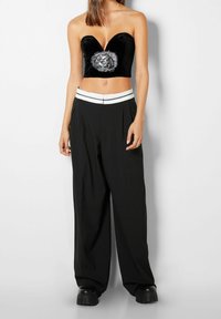 Musta samettinen olkaimeton toppi, syvä V-pääntie ja paljettikoristelu, yhdistettynä löysiin mustiin housuihin, joissa on valkoinen vyötärönauha.