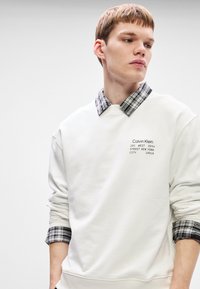 Λευκή μπλούζα με κουκούλα, με μια μαύρη και γκρίζα πουκαμίσα με καρώ σχέδιο από κάτω. Διαθέτει λογότυπο Calvin Klein στο στήθος και ραμμένες μανσέτες.