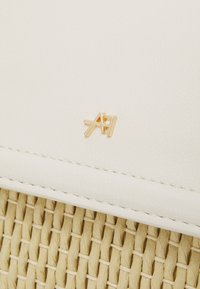 Malé zlaté kovové logo se vzájemně propojenými písmeny "AH" na hladké bílé kůži nad tkaným béžovým materiálem.