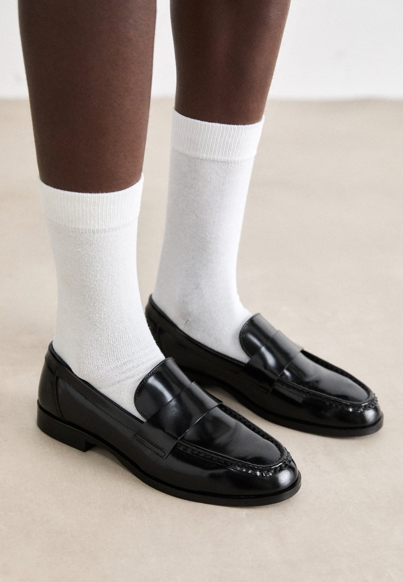 Mocassini in pelle verniciata nera con un design cucito, abbinati a calzini di cotone bianchi a coste, mostrati su uno sfondo di pavimento chiaro.