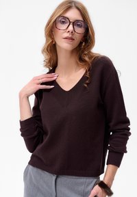 Kobieta z falującymi, jasnobrązowymi włosami, w dużych okrągłych okularach w stylu żółwia, ciemnofioletowym swetrze z dekoltem w serek, szarych spodniach i futrzanej bransoletce, pozująca.