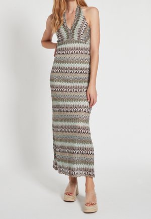 Mujer con vestido maxi sin mangas de estampado y bordes ondulados, y sandalias de plataforma transparentes, de pie contra un fondo liso.