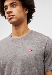 Camiseta de algodón gris con cuello redondo y mangas cortas, que presenta un pequeño parche rojo del logo de Levi's en el lado izquierdo del pecho.