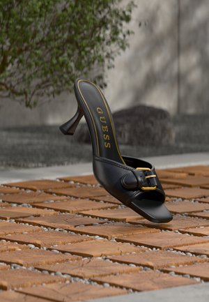 Mula negra de tacón alto con hebilla dorada y logo "GUESS" en el interior, situada sobre un suelo de baldosas exteriores con textura y fondo desenfocado de vegetación.