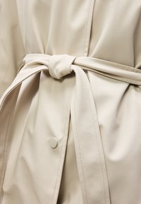 Μπεζ παλτό τρεντς δεμένο στη μέση με φαρδιά ζώνη και με ένα κουμπί στο μπροστινό μέρος.