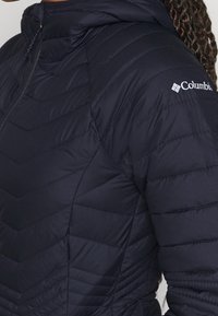 Närbild av en person som bär en svart vadderad Columbia-jacka med huva och logotyp på överärmen.