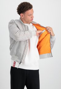 Šedá bomber bunda s rebrovanými manžetami, prešívaným povrchom a oranžovým vnútrom. Model otvára bundu a odhaľuje vnútornú kapsu. Pod ňou biela mikina.