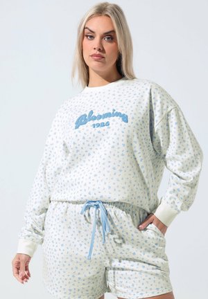 Mujer rubia con sudadera blanca y pantalones cortos con pequeño estampado floral azul y texto "Blooming 1986" en la sudadera.