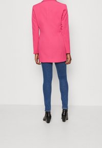 Veste blazer rose cintrée à manches longues, col cranté et silhouette ajustée, associée à un jean skinny bleu foncé et des bottines noires.