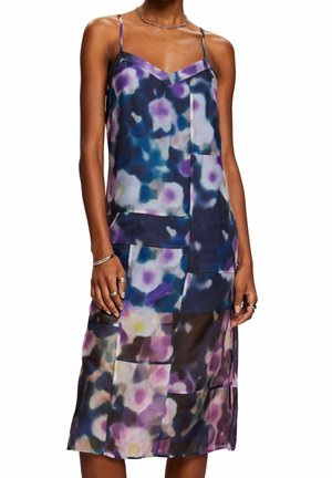 Femme portant une robe midi sans manches avec de fines bretelles et un motif floral abstrait coloré dans des tons de bleu, violet et blanc.