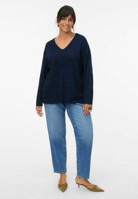 Maglione lavorato a maglia blu navy con scollo a V; vestibilità ampia e maniche lunghe abbinato a jeans a gamba dritta blu chiaro e scarpe verdi con punta a stiletto.