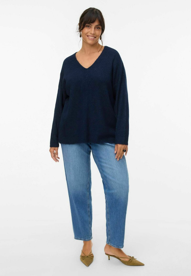 Maglione lavorato a maglia blu navy con scollo a V; vestibilità ampia e maniche lunghe abbinato a jeans a gamba dritta blu chiaro e scarpe verdi con punta a stiletto.