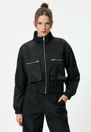 Schwarze, kurz geschnittene Jacke aus glattem, reflektierendem Gewebe. Verfügt über einen hohen Kragen, zwei mit Reißverschluss verschließbare Brusttaschen und elastische Bündchen.