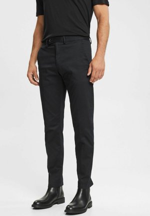 Uomo con pantaloni neri aderenti, maglietta nera e stivali di pelle neri, in piedi davanti a uno sfondo chiaro e uniforme.