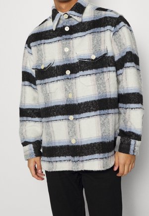 Homme portant une veste-chemise en laine à carreaux boutonnée avec des rayures horizontales noires, grises et bleues et des poches poitrine à rabat.