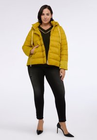 Žlutá péřová bunda s kapucí, s diagonálně prošívanými sekcemi, dvěma předními kapsami a předním zipem. Nositelná přes tmavou svetr a černé džíny.