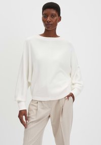 Maglione bianco e morbido con scollatura ampia e spalle scese. Abbinato a pantaloni beige chiaro plissettati. Texture liscia, vestibilità rilassata.