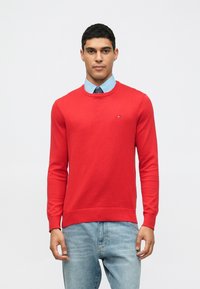 Άνδρας φορεμένος με κόκκινο πουλόβερ τύπου crewneck πάνω από ανοιχτό μπλε πουκάμισο και σκούρη γραβάτα, συνδυασμένος με ανοιχτό μπλε τζιν, στέκεται μπροστά σε λευκό φόντο.