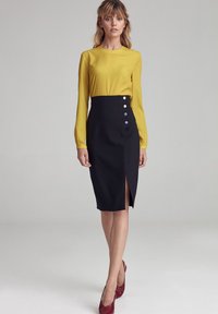 Żółta bluzka z długim rękawem w Połączeniu z dopasowaną czarną spódnicą z przednim rozcięciem i trzema kolorowymi guzikami po boku.