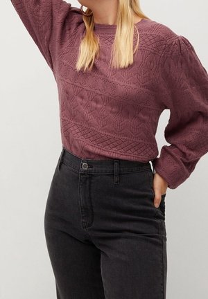 Vrouw draagt een gestructureerde mauve gebreide trui en hooggetailleerde zwarte spijkerbroek, poseert met één arm omhoog en de andere hand in de zak.