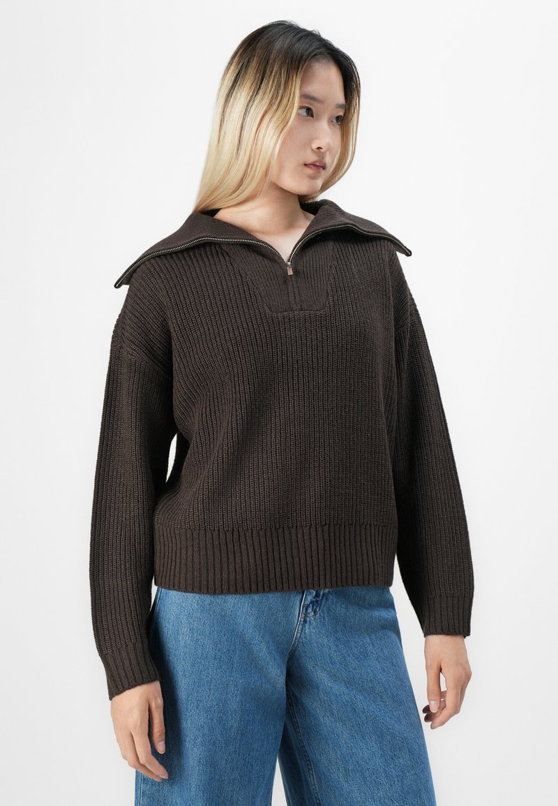 Femme jeune avec de longs cheveux ombrés portant un pull en laine brun foncé avec un col zippé et un jean en denim bleu, se tenant devant un fond blanc.