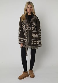 Vrouw in een bruin met patronen versierde fringes cardigan, zwarte leggings en beige instappers voor de winter, staande tegen een eenvoudige achtergrond.