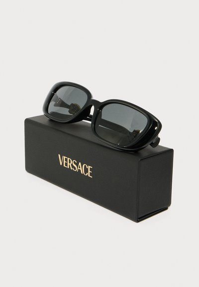 Černé oversized sluneční brýle s hladkým, lesklým rámem, tmavými čočkami a zlatými akcenty na stranici, prezentované v černém pouzdře Versace.