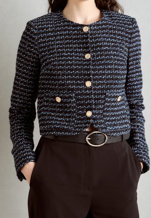 Femme portant une veste boutonnée texturée noire et bleue avec des boutons dorés et un pantalon noir avec une ceinture noire avec une boucle ovale argentée.